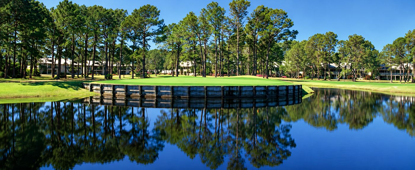 Island green ringed by a wooden bulkhead, reflected in a still lake under tall pines