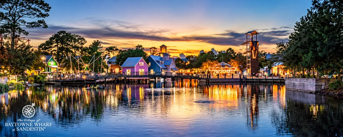 Colorful village shops and an adventure tower lit at sunset, reflected in a pond at Baytowne Wharf