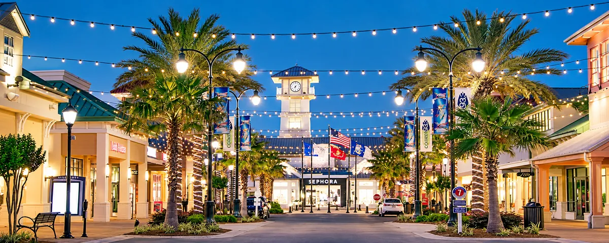 Palm-lined shopping street with string lights and a clock tower at dusk
